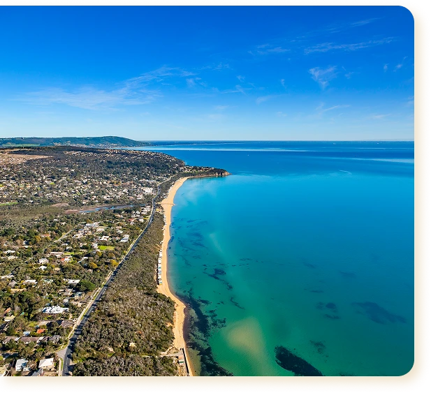 A panoramic aerial photograph of the coastline on a bright, sunny day. The image captures a bird’s-eye view of the endless bright-blue sea, which gives way to turquoise shallows near the shore, and a narrow strip of sandy beach. A dense green forest stretches along the coast, behind which lies a coastal residential area with detached houses and roads leading off into the distance towards the hills on the horizon. The atmosphere is serene, summery and spacious.